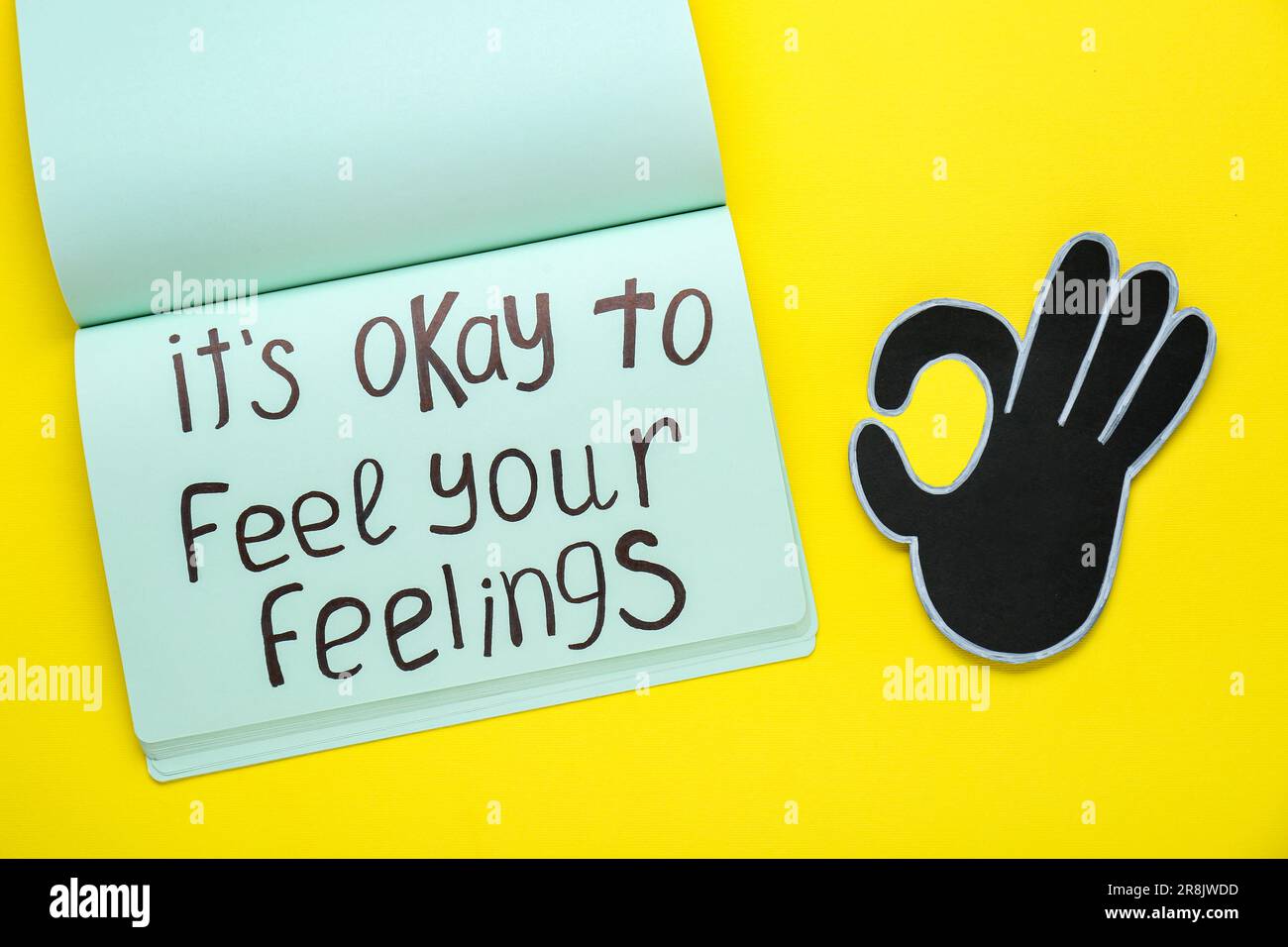 Notebook with phrase It`s Okay To Feel Your Feelings and paper cutout of OK hand gesture on yellow background, top view Stock Photo